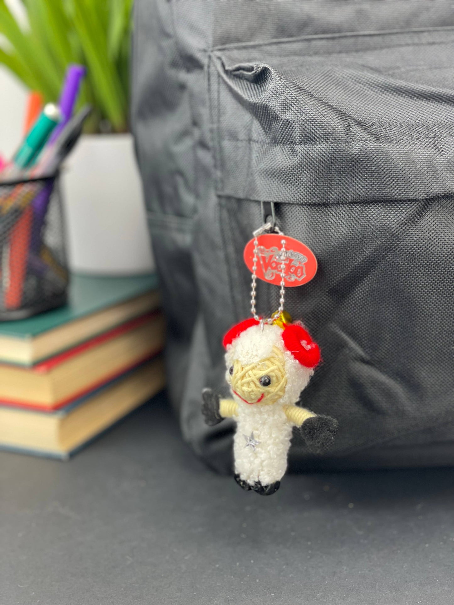 Small crocheted sheep keychain with red accents on a gray surface, with books and a pen holder in the background.