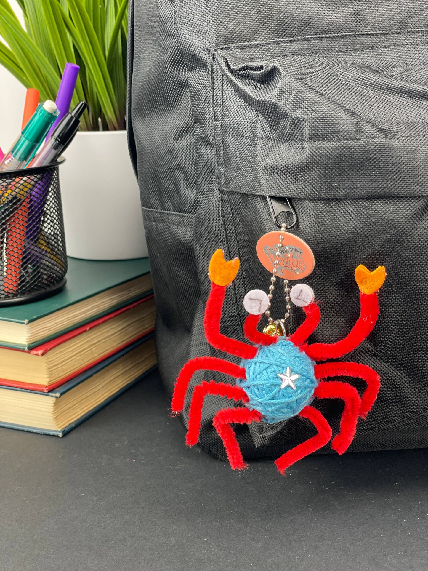 Colorful crab-shaped keychain attached to a backpack with stationery items in the background.