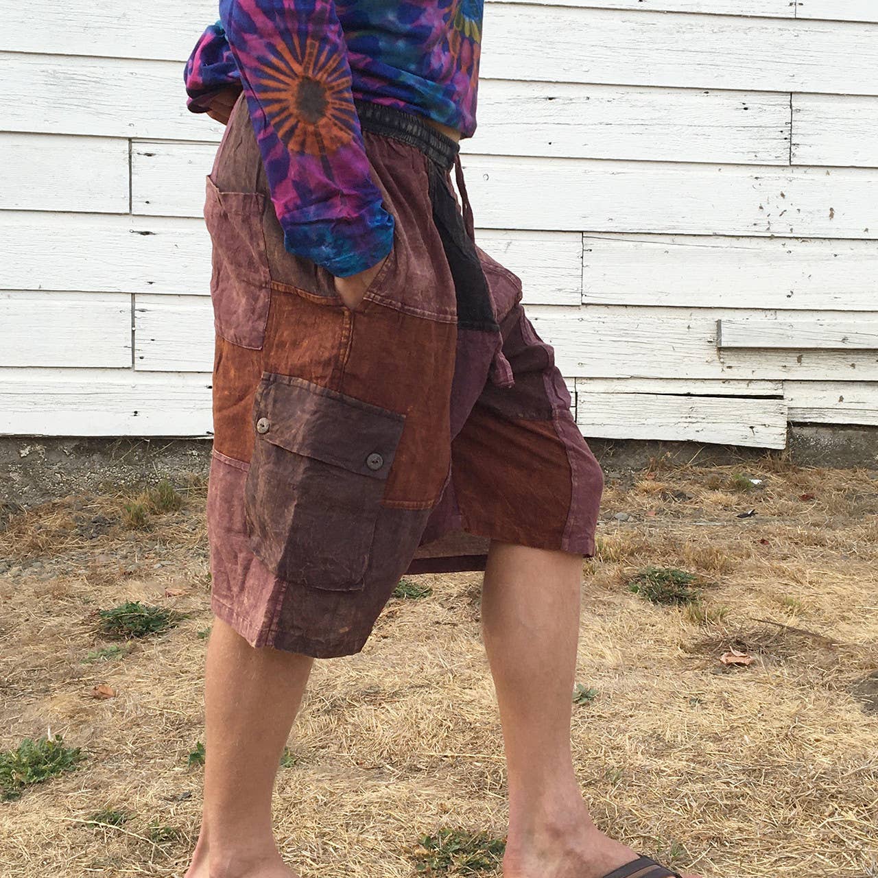 Person wearing a tie-dye shirt and brown shorts standing in front of a wooden wall.