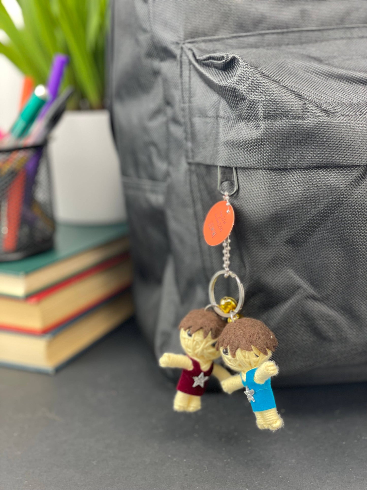 Two small figurines on a keychain attached to a backpack with books and stationery items in the background.