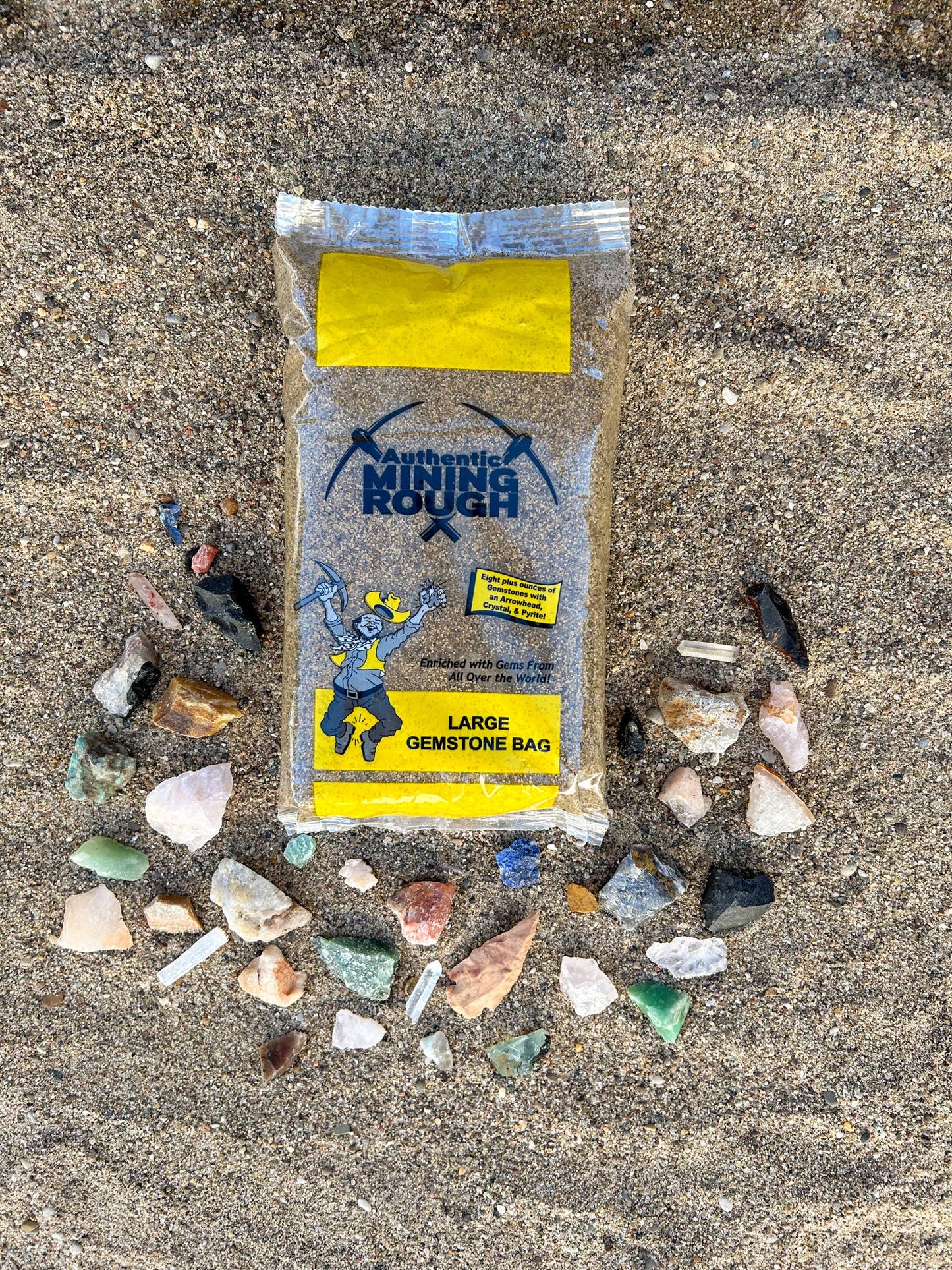 Package of 'Authentic Mining Rough' gemstone bag on a sandy surface with various stones around it.