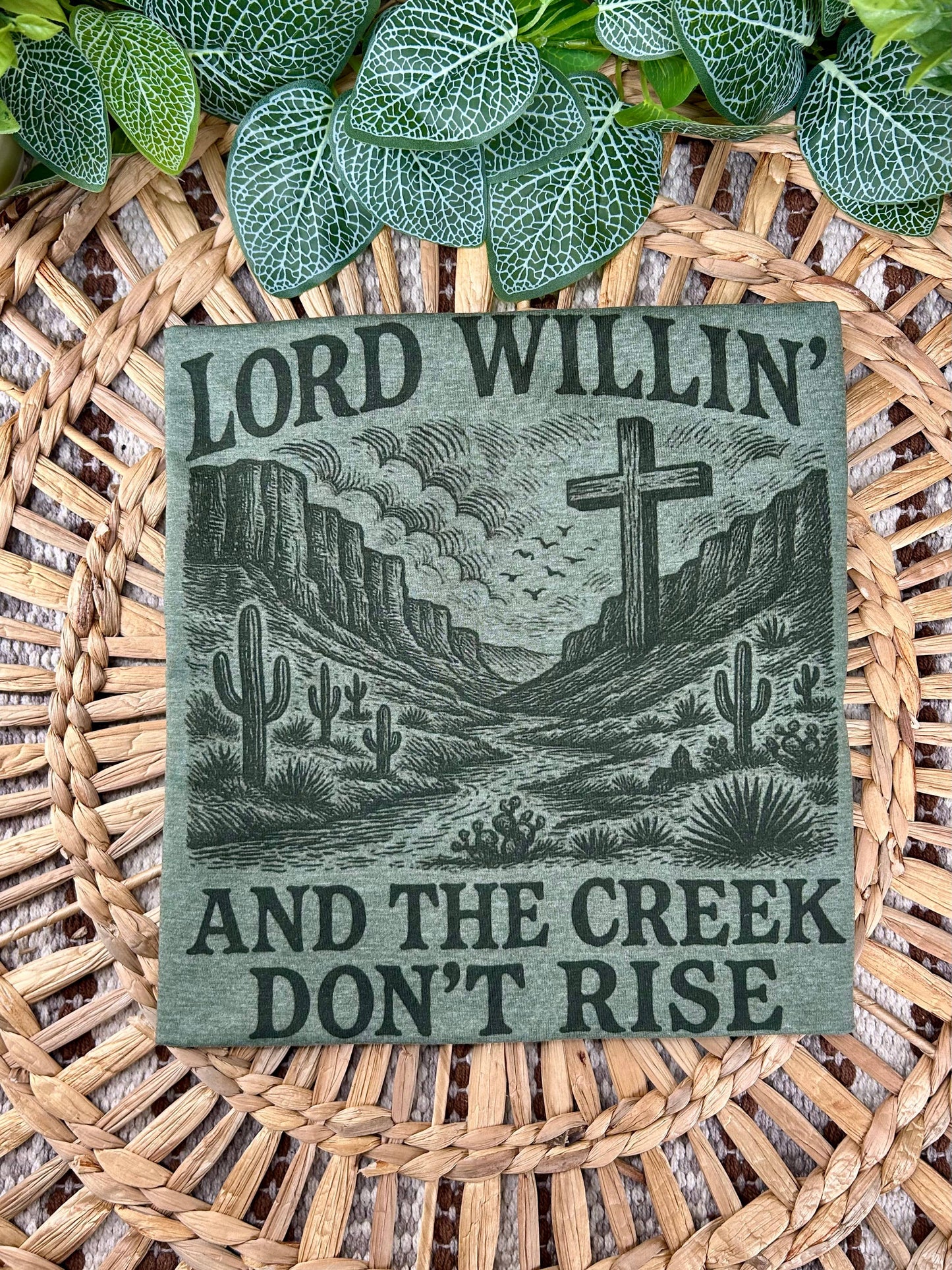 Decorative sign with desert landscape and cross, featuring the text 'Lord Willin' and the Creek Don't Rise', on a woven surface.