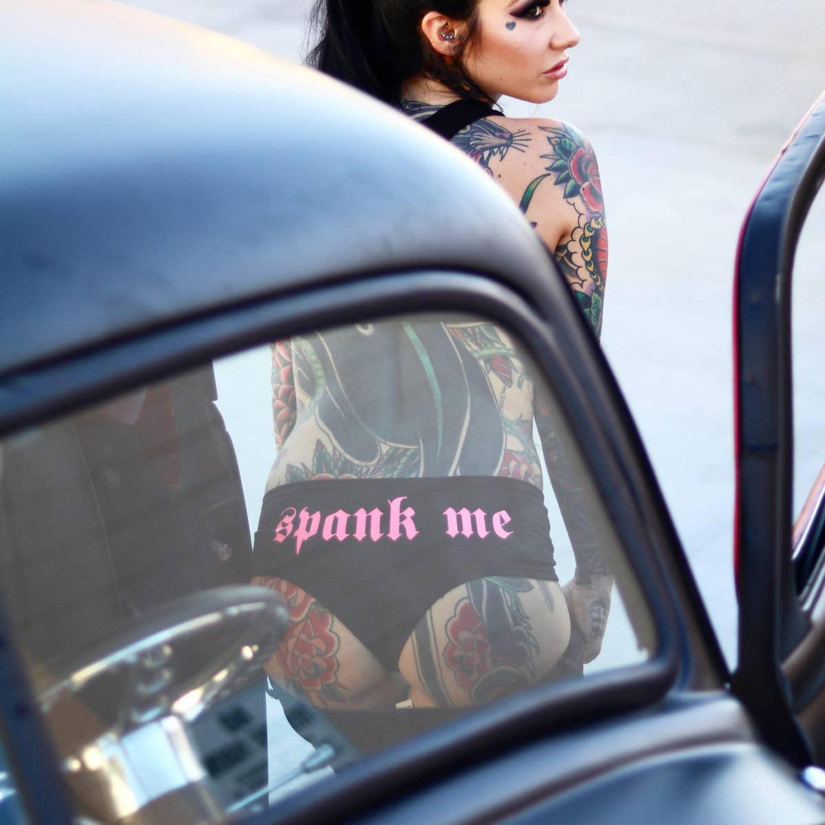 Person with tattoos wearing a black thong with 'spank me' text, reflected in a car mirror.