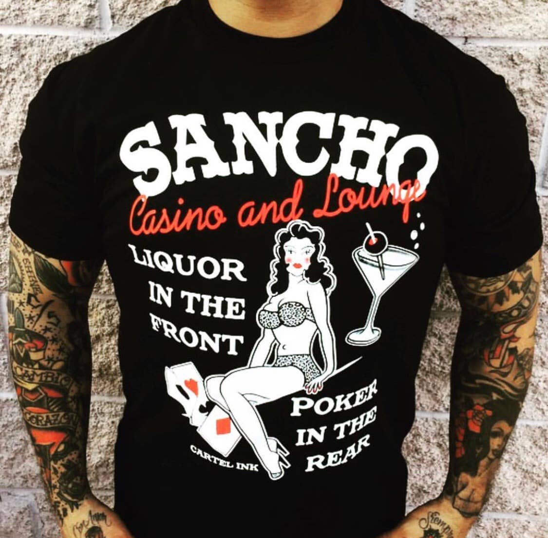 Black t-shirt with a graphic design featuring a woman, cocktail, and text on a brick wall background.