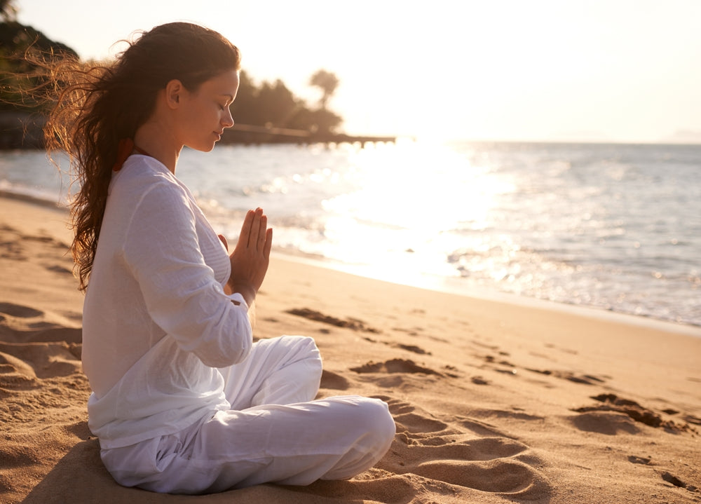 Beautiful woman in white clothing sitting on beach sand meditating. 