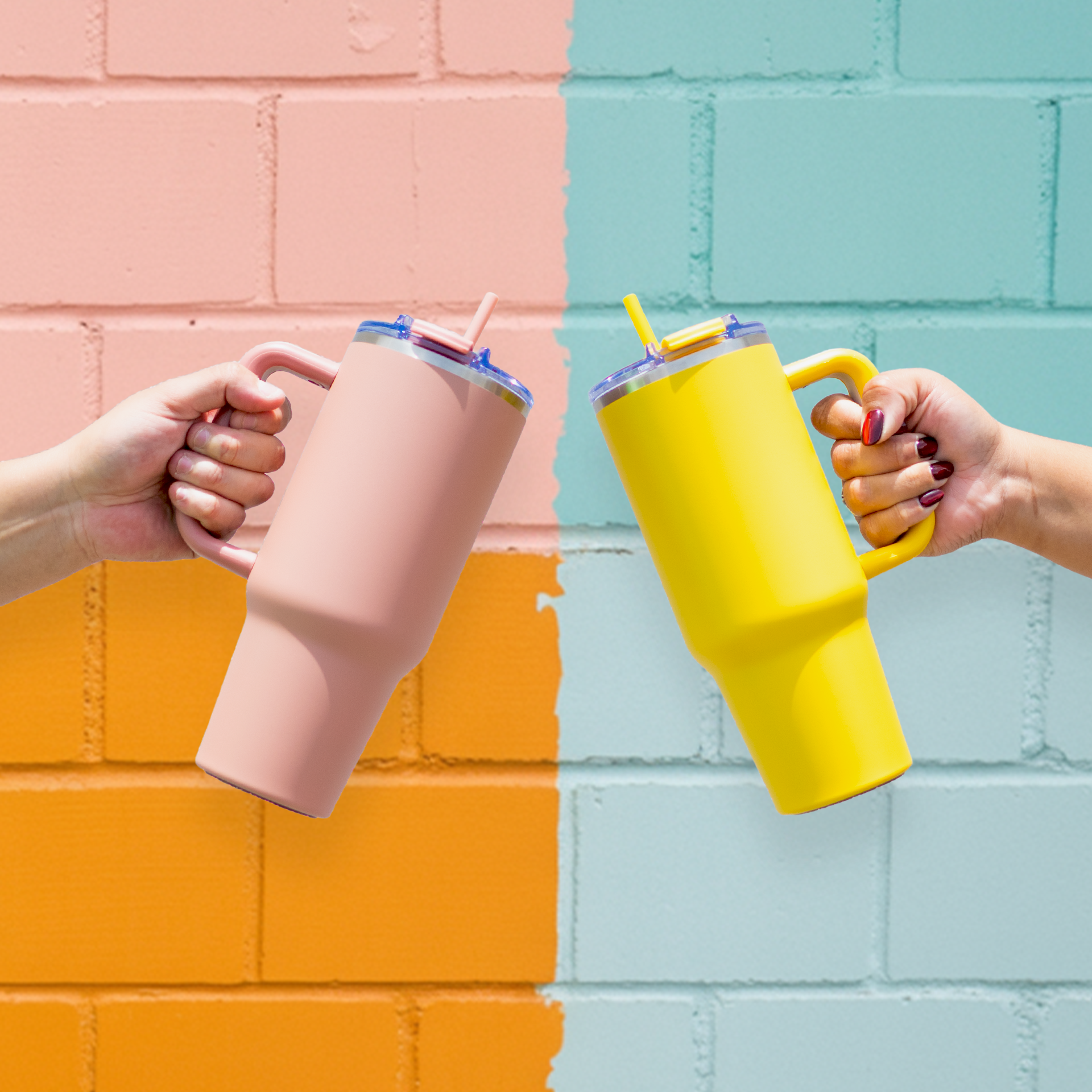 Two hands holding pink and yellow tumblers against a colorful brick wall.