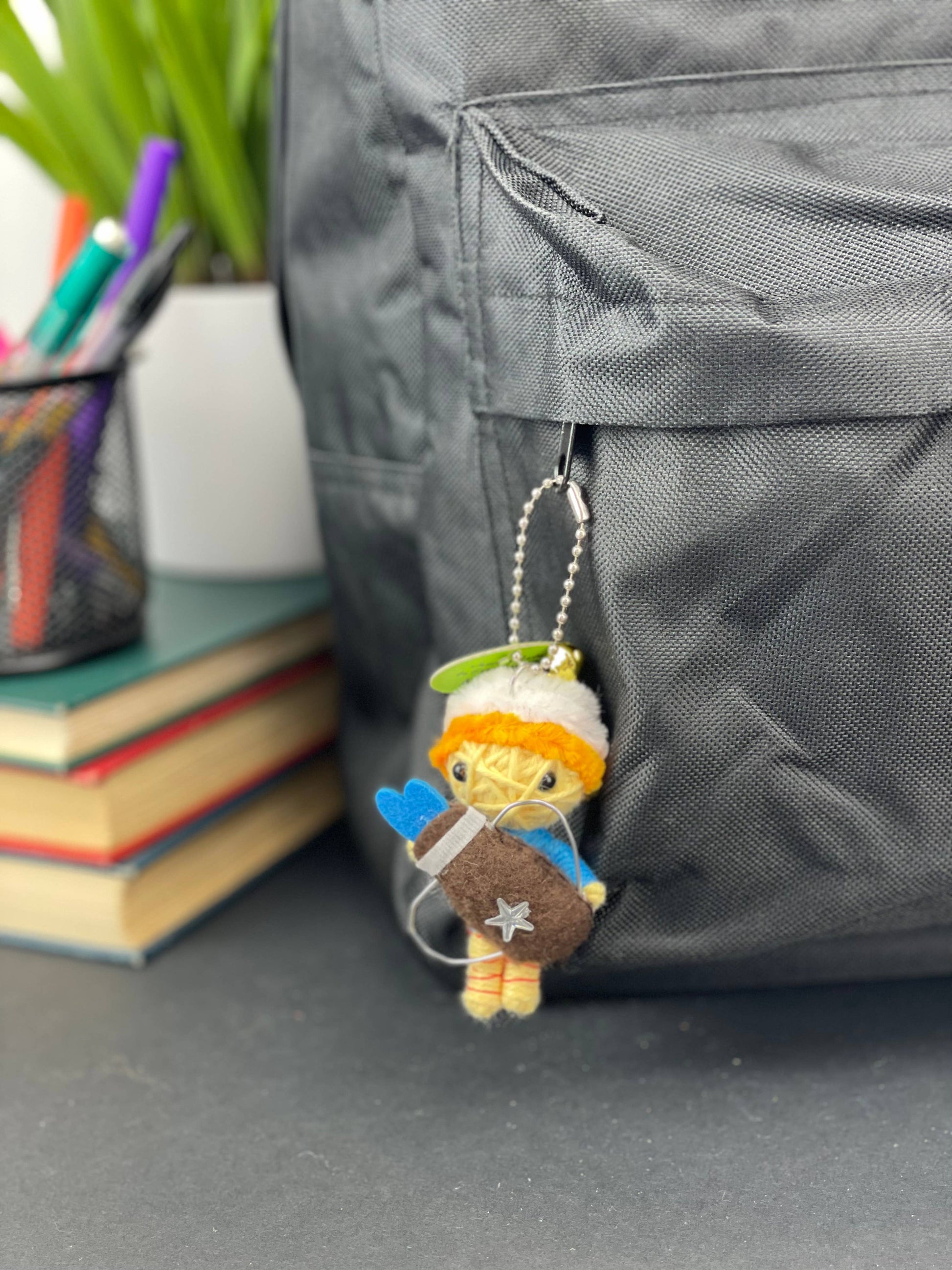 Small figurine keychain attached to a backpack with books and stationery items in the background.