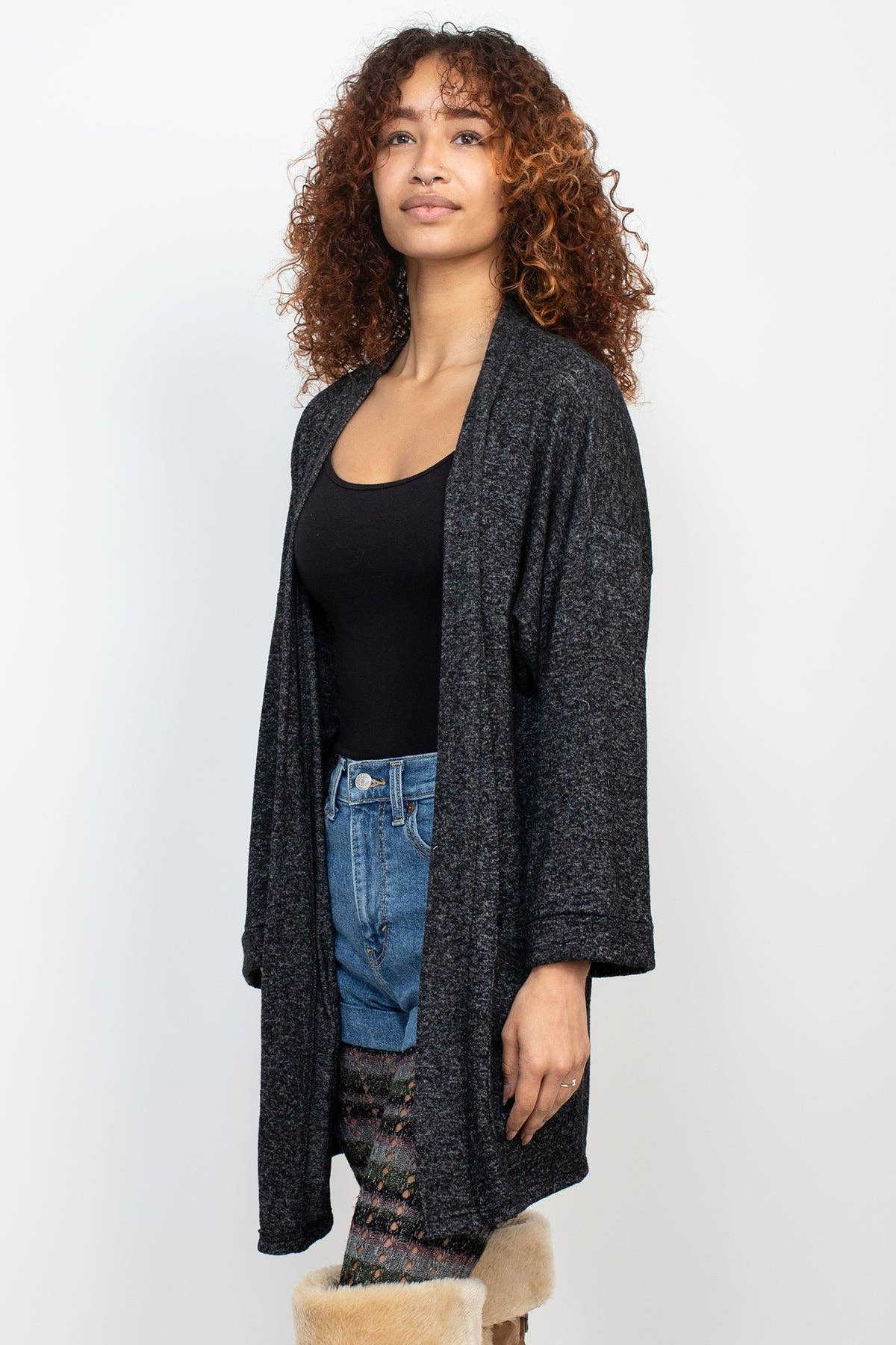 Woman wearing a long dark gray cardigan over a black top and blue jeans on a white background