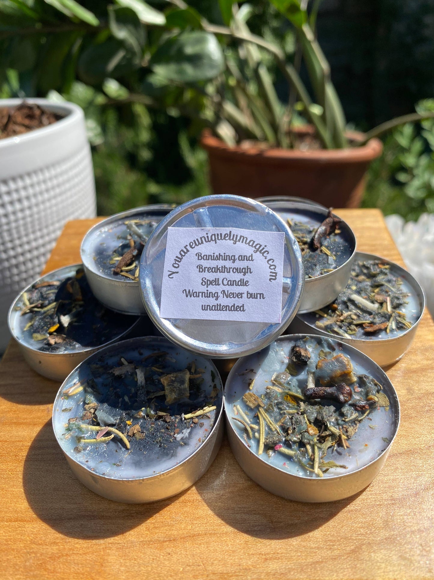 Collection of small metal containers with candle-like contents on a wooden surface, surrounded by plants.
