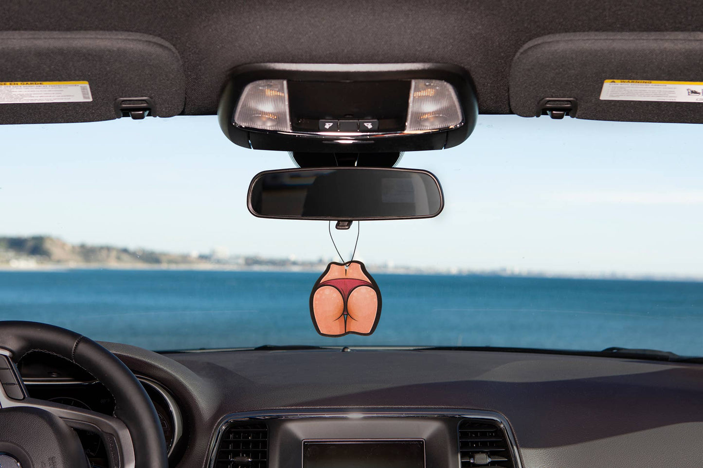 Car air freshener shaped like a butt hanging from a car's rearview mirror with a scenic background.