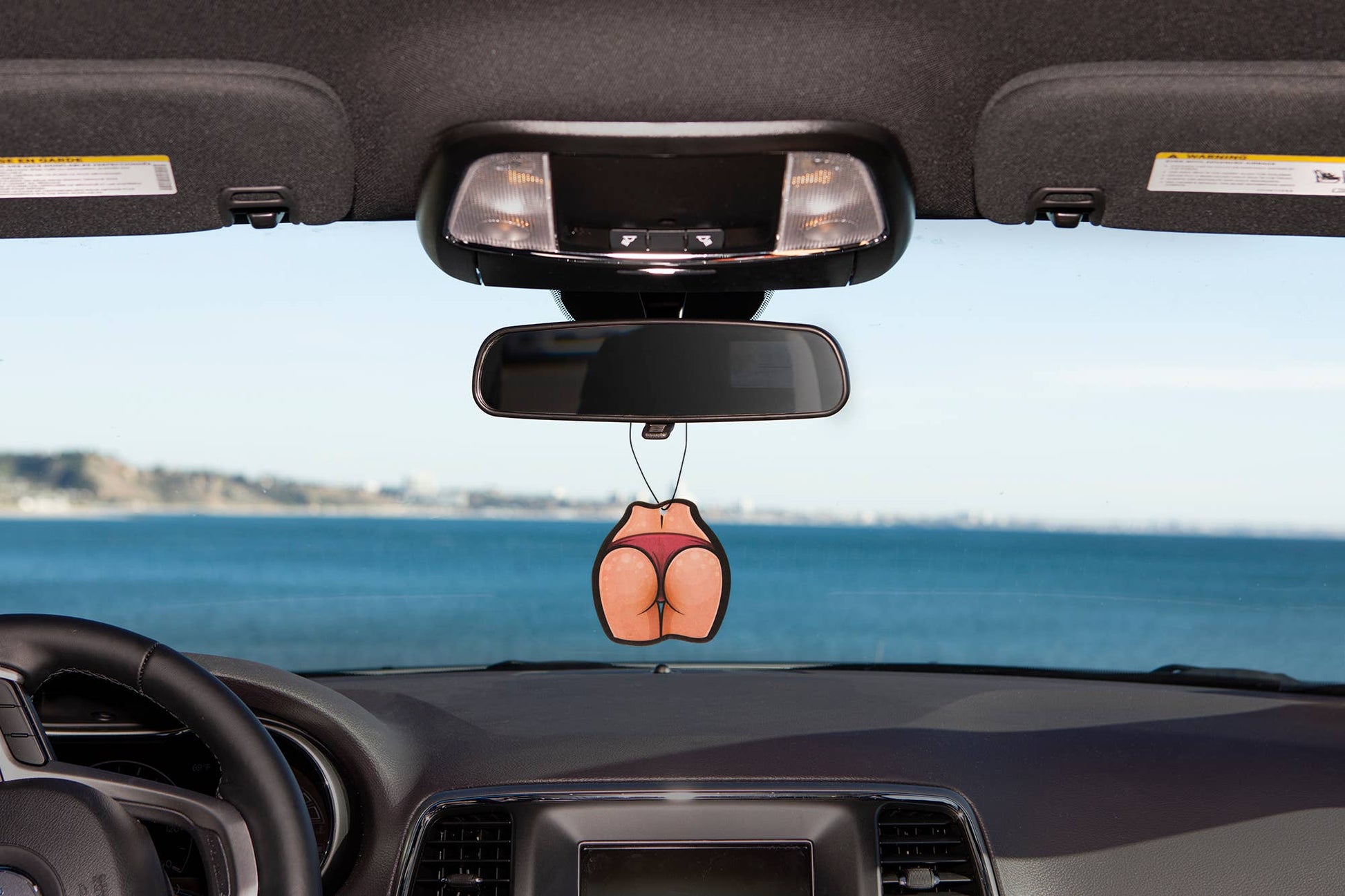 Car air freshener shaped like a butt hanging from a car's rearview mirror with a scenic background.