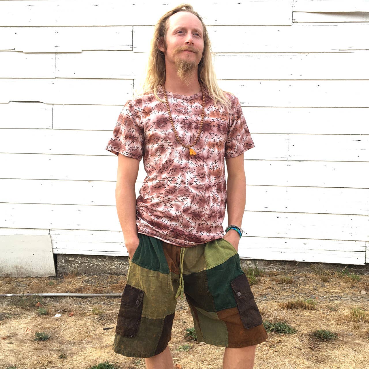 Man wearing a tie-dye shirt and patchwork shorts standing against a white wooden wall.