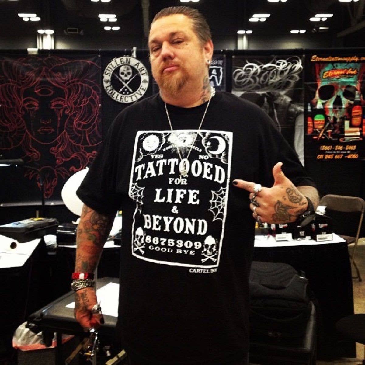 Man wearing a black t-shirt with a tattoo-themed design in a tattoo studio setting.