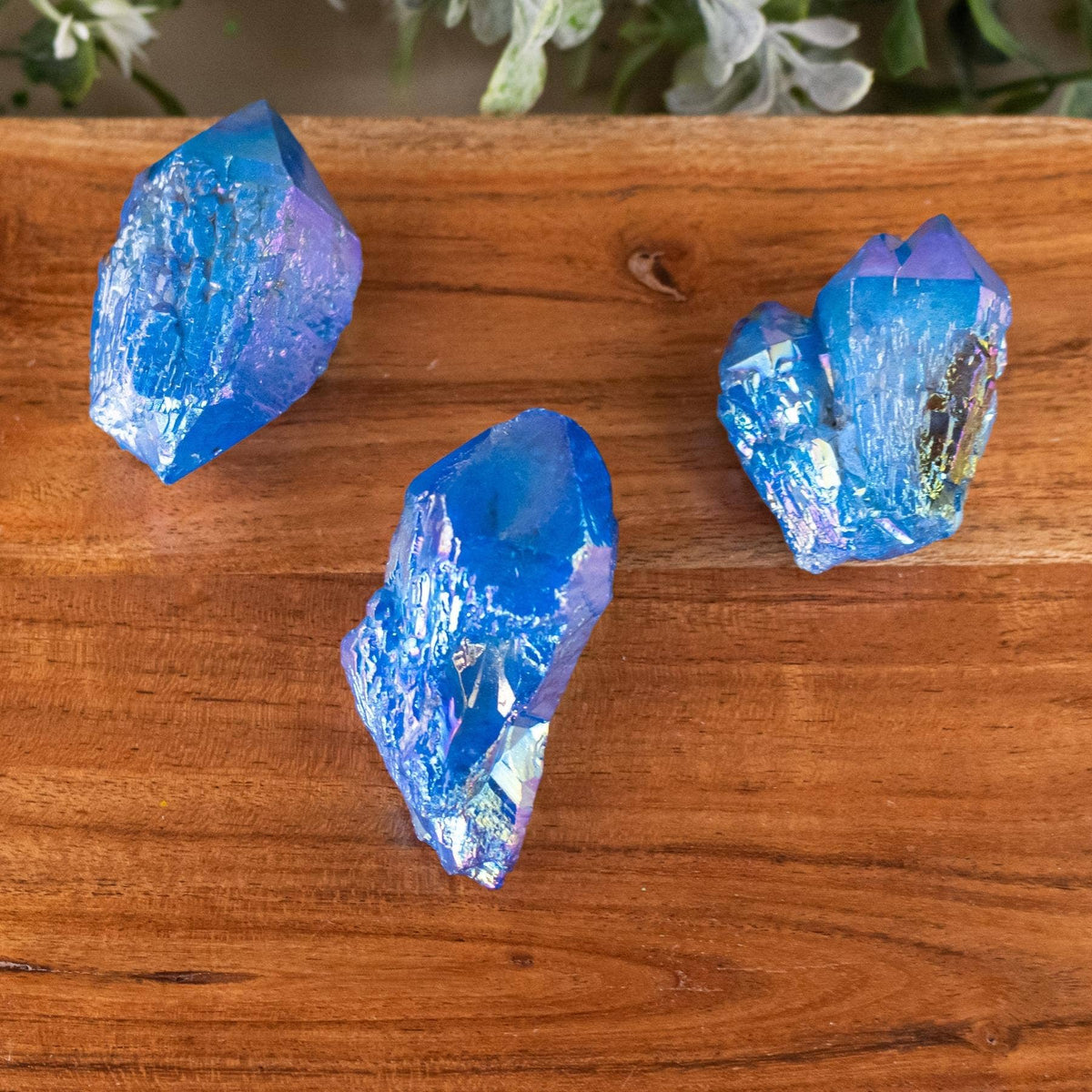 Three blue iridescent crystal points displayed on a wooden surface with soft green foliage blurred in the background.