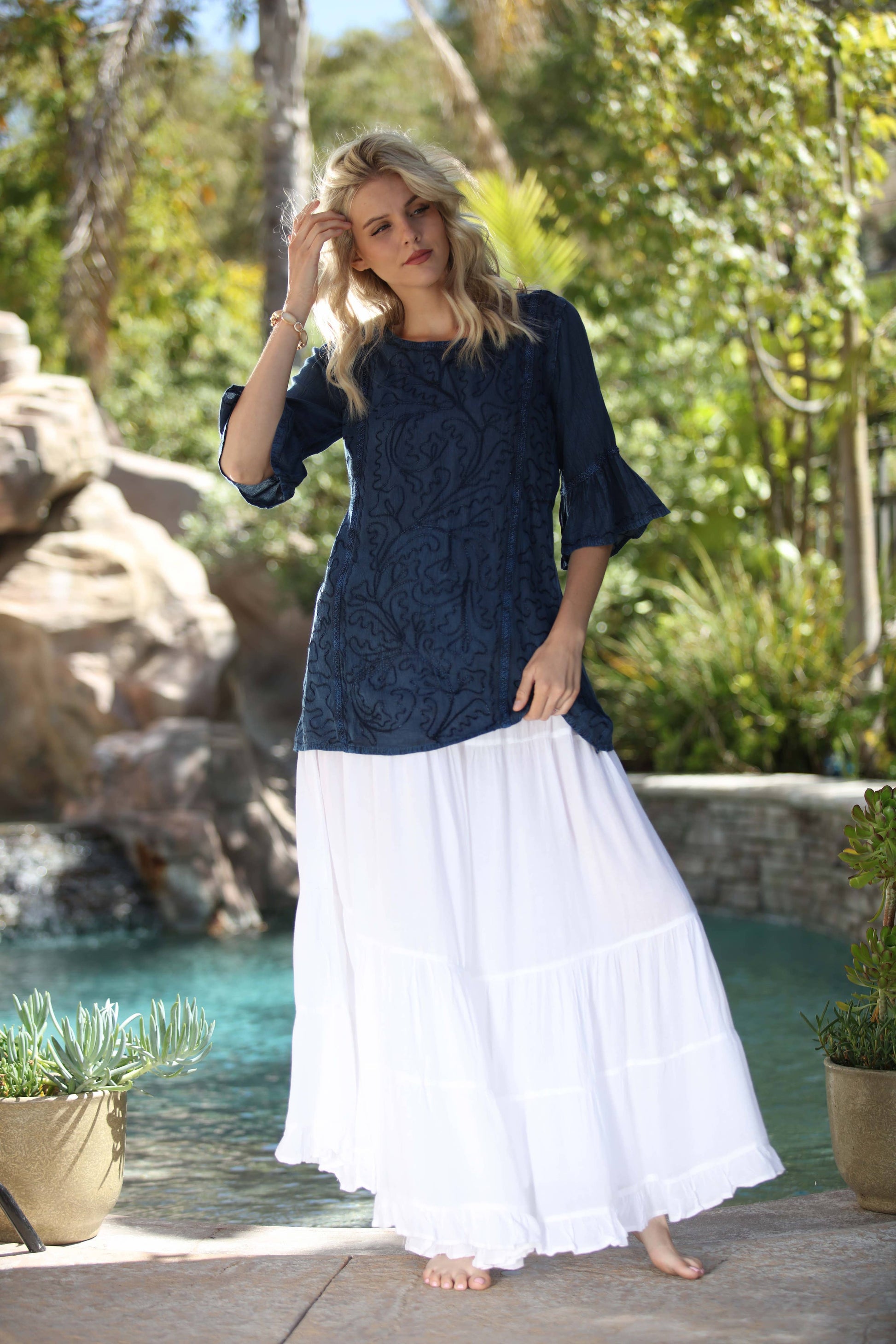 Woman wearing a navy top and white skirt standing by a pool with greenery in the background