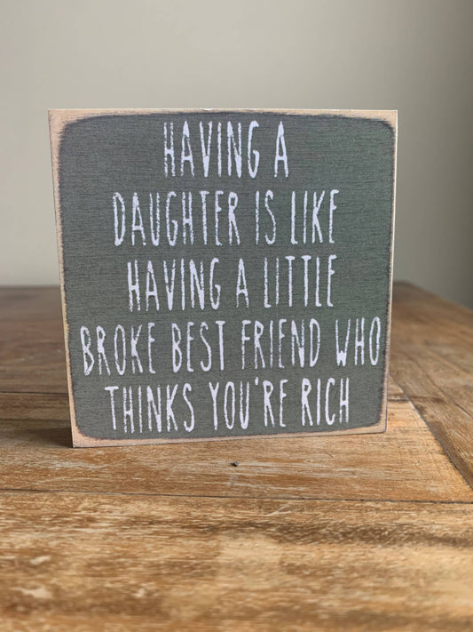 Wooden sign with humorous quote about having a daughter on a wooden surface.