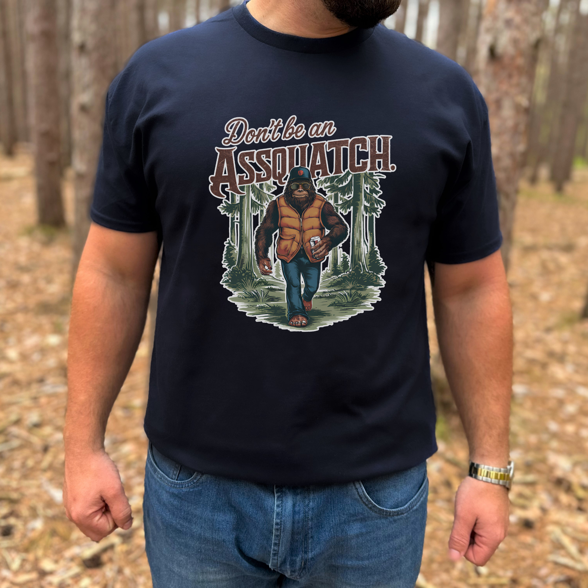 Person wearing a navy blue t-shirt with a graphic design of a person in a forest and text 'Don't be an Assquatch'.