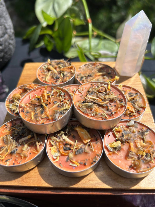 Small candles with dried herbs on a wooden board, surrounded by greenery and a crystal.