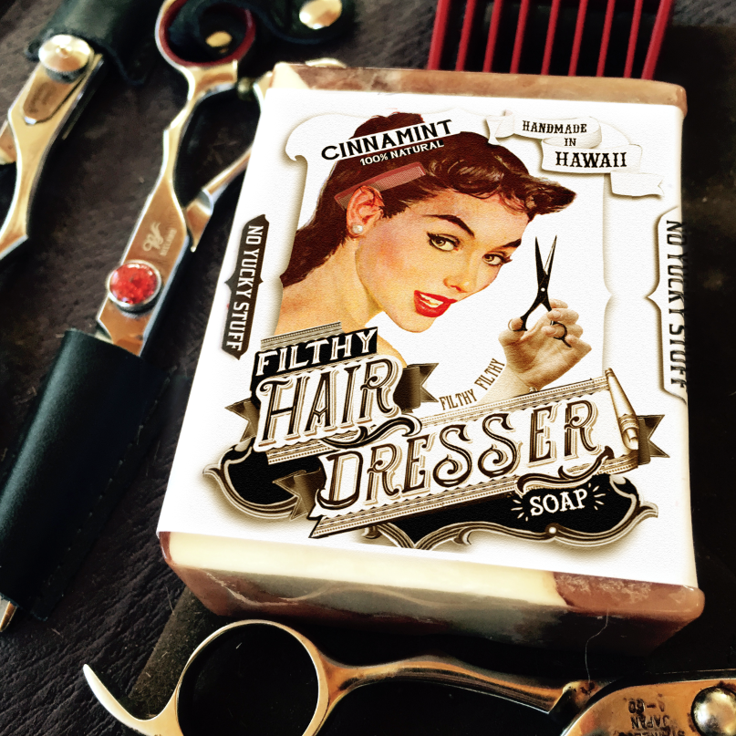 Bar of soap with a vintage-style label featuring a woman and text, surrounded by hairdressing tools on a dark surface.