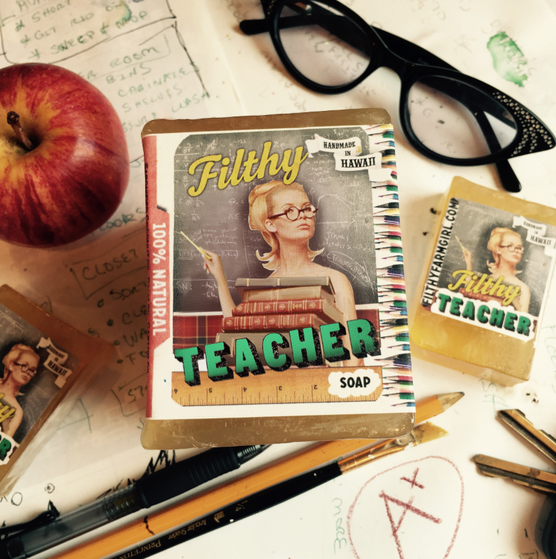 Soap packaging labeled 'Filthy Teacher' on a desk with an apple, glasses, and stationery items.