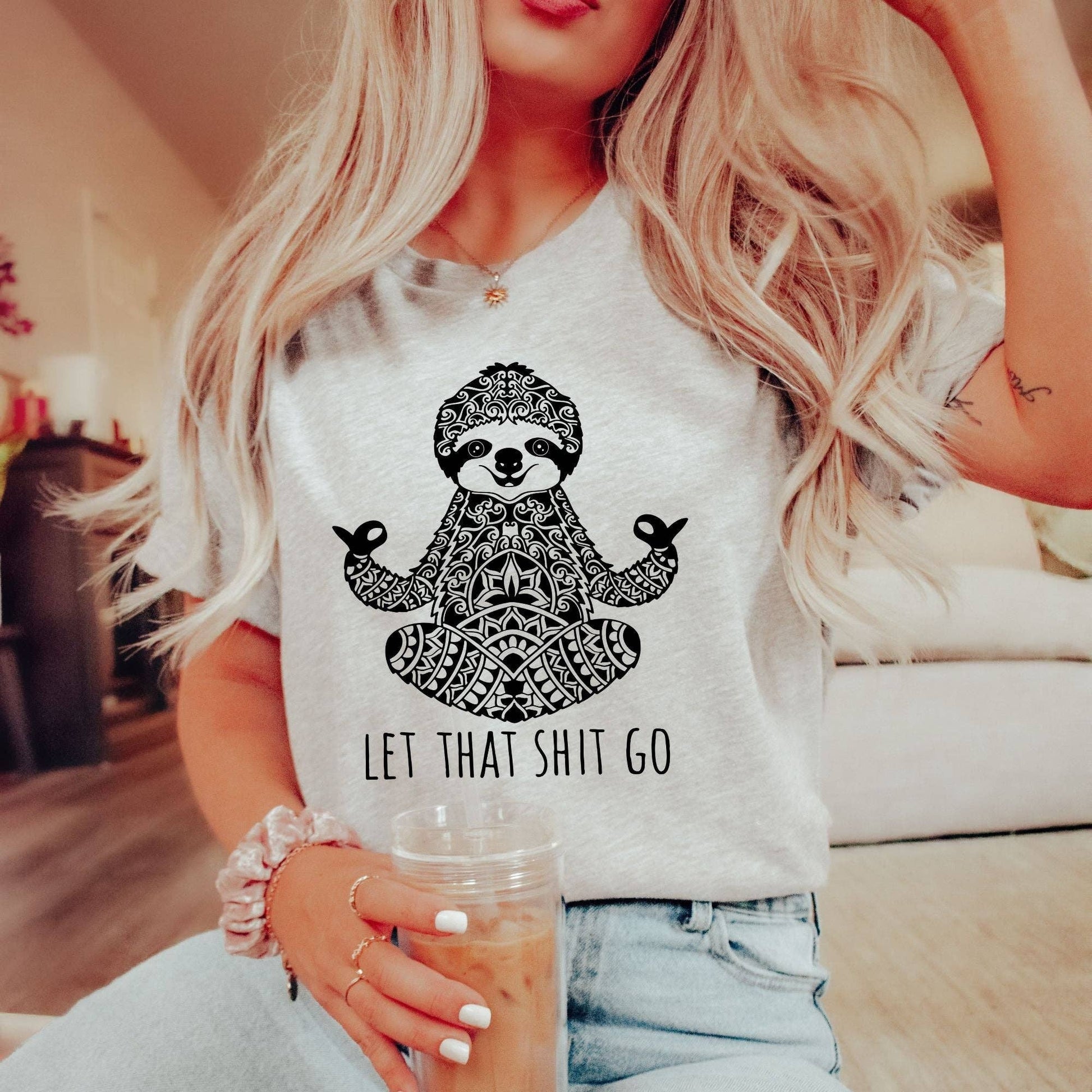 Person wearing a white t-shirt with a sloth graphic and text, holding a drink.