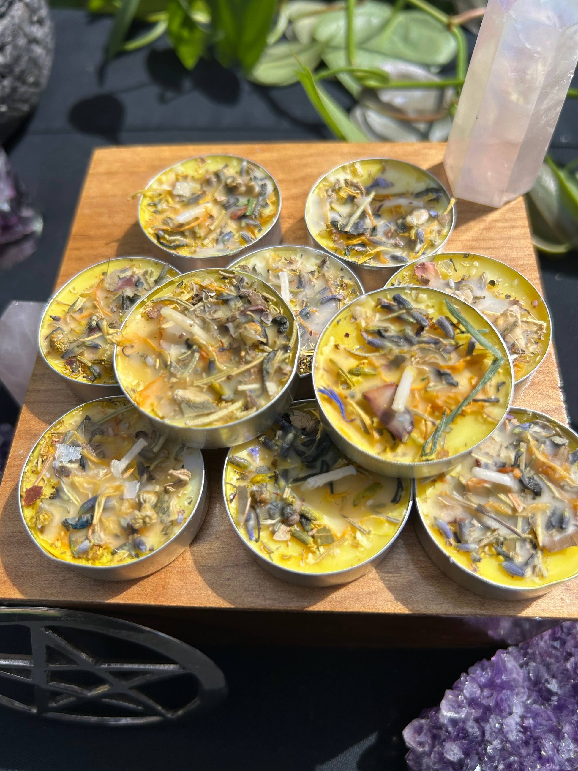 Small candles with decorative elements on a wooden board, surrounded by crystals and plants.