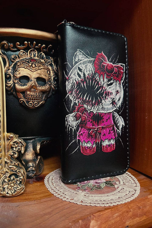 A black leather zip-around wallet featuring a gothic horror graphic of a cat with sharp teeth, a red bow, and pink shorts, displayed on a wooden shelf next to decorative skull masks.
