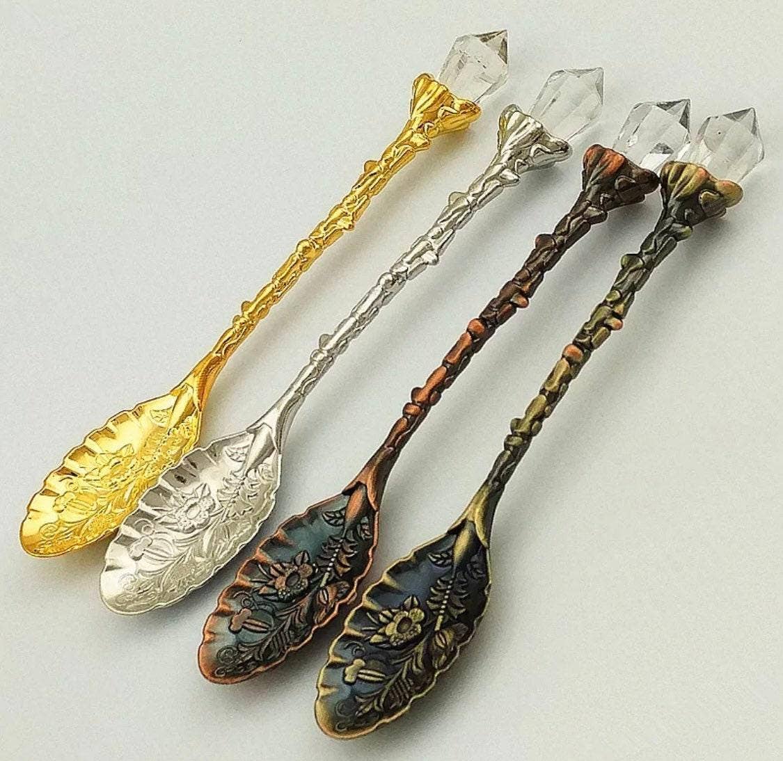 Four decorative spoons in gold, silver, bronze, and green with crystal handles on a light gray background.