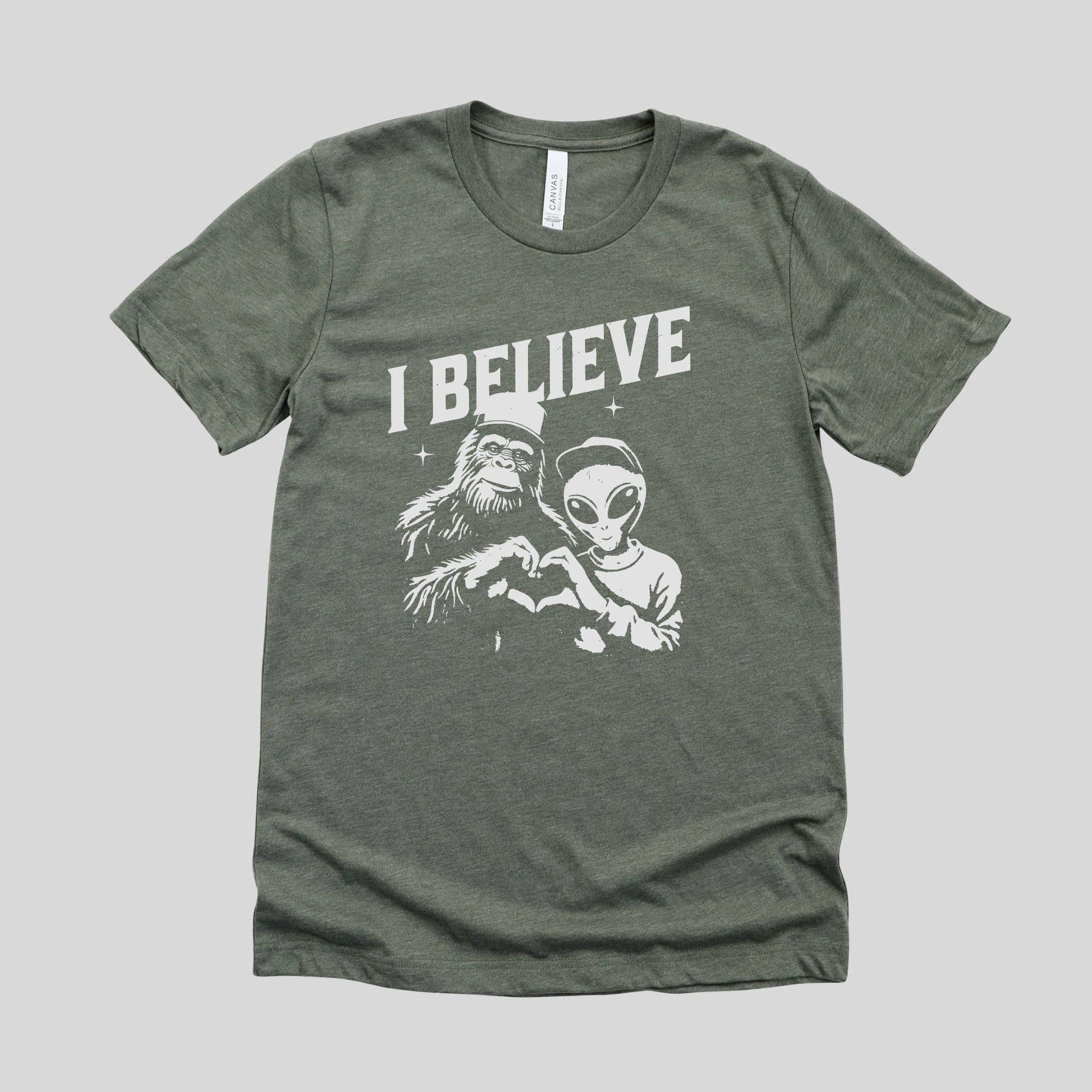 Green t-shirt with 'I Believe' graphic featuring a caveman and alien on a gray background