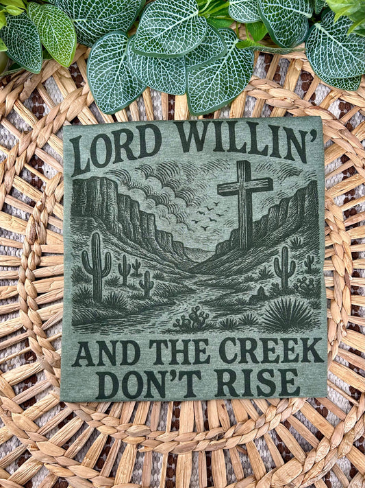 Decorative sign with desert landscape and cross, featuring the text 'Lord Willin' and the Creek Don't Rise', on a woven surface.
