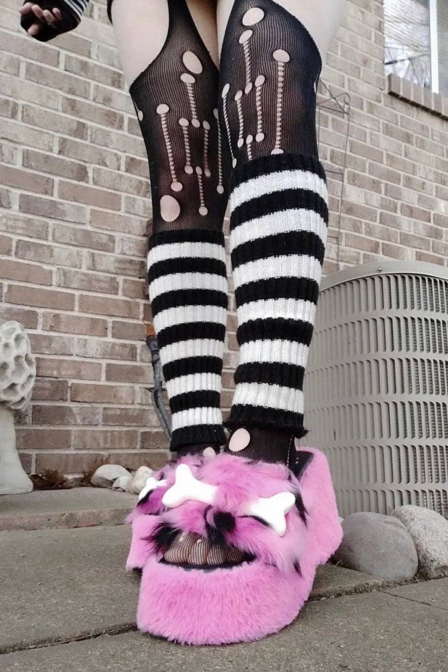 Person wearing black and white patterned tights with pink fuzzy slippers in front of a brick wall.