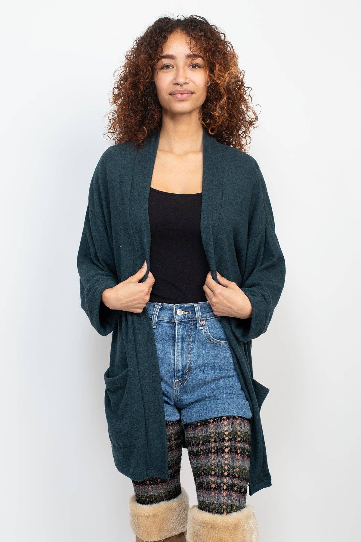Woman wearing a teal cardigan, black top, blue jeans, and patterned leggings on a white background