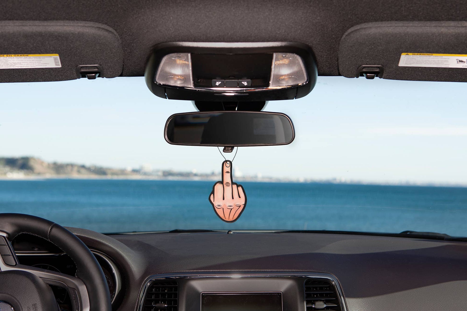 Car dashboard with a humorous rearview mirror accessory showing a middle finger, set against a scenic background.