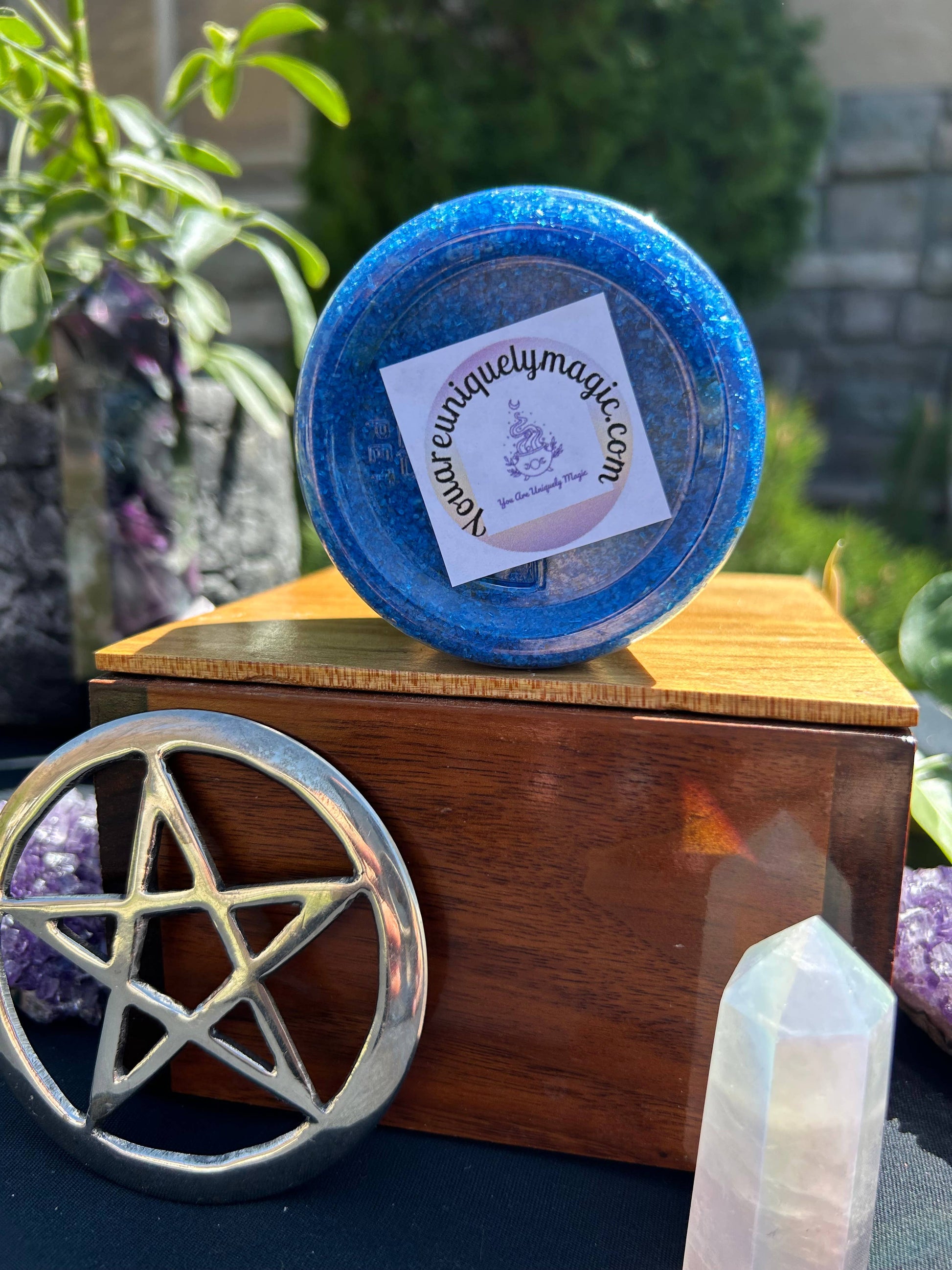 Blue container with a label on top of a wooden box with a pentagram symbol, surrounded by crystals and plants.