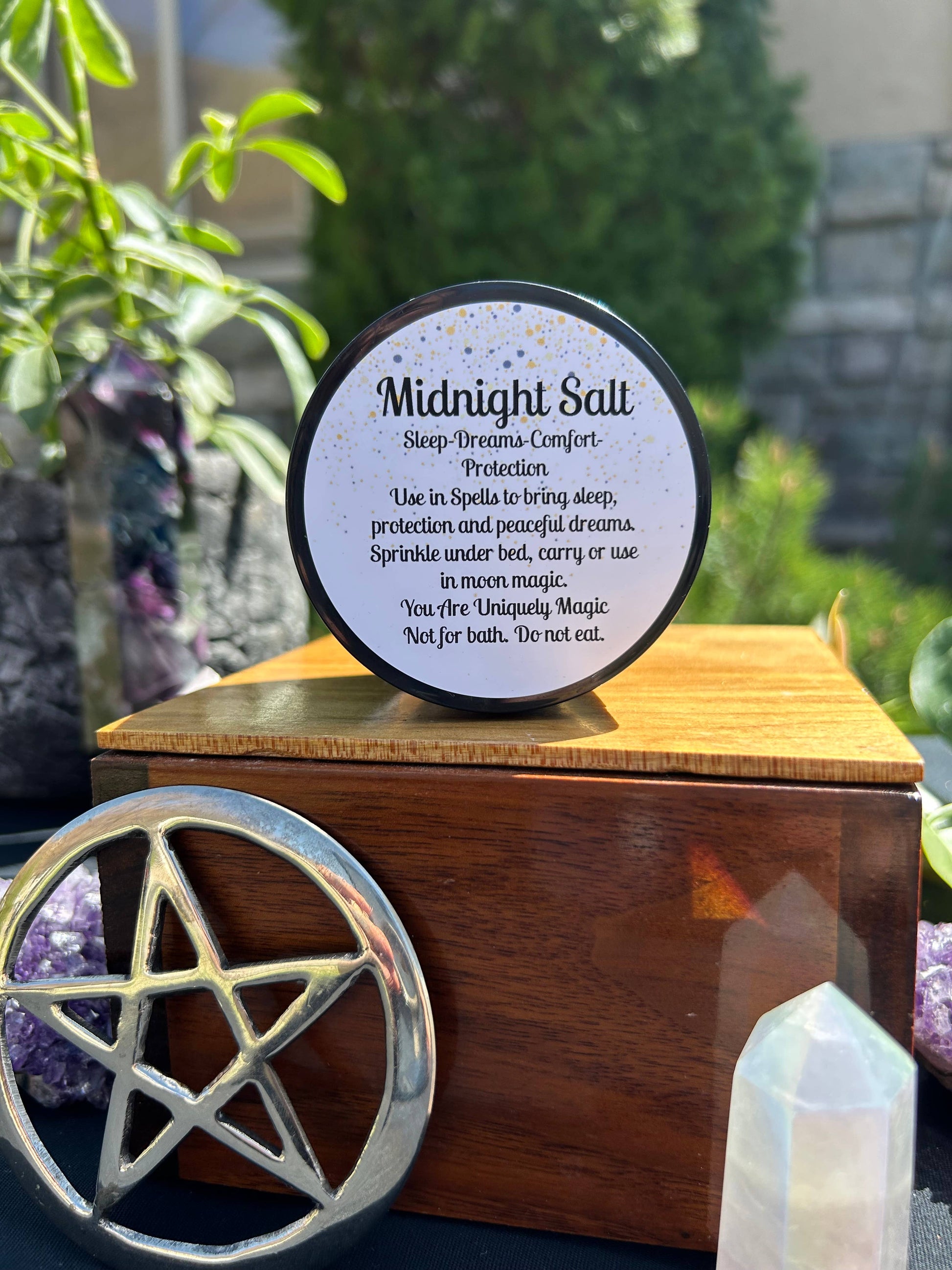 Midnight Salt container on a wooden stand with a pentagram symbol, surrounded by plants and crystals outdoors.