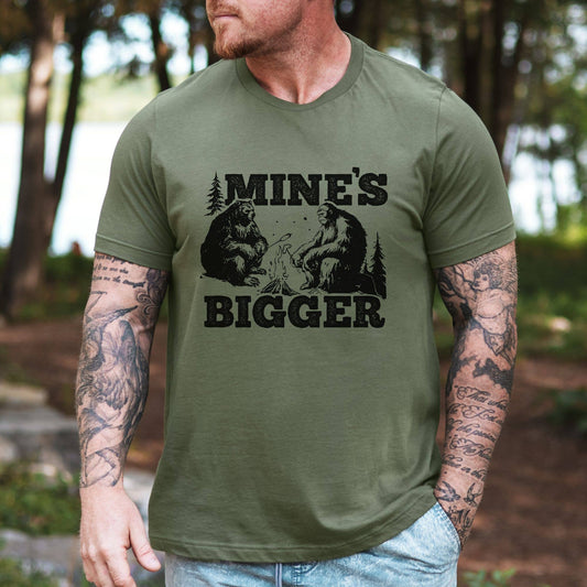 Man wearing a green t-shirt with 'MINE'S BIGGER' graphic and bear design outdoors.