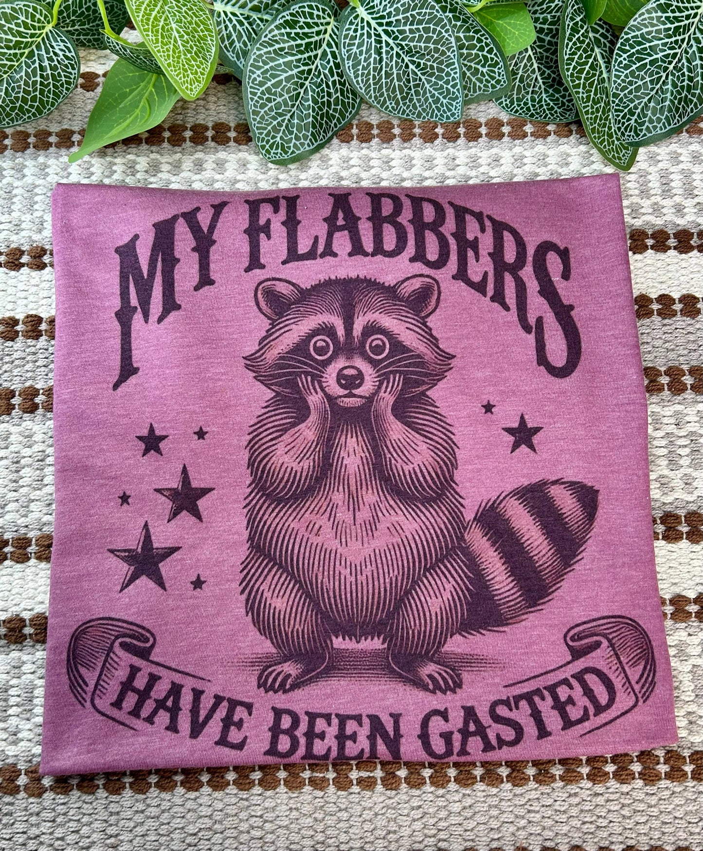 Pink towel with raccoon illustration and text 'My flabbers have been gasted' on a textured surface with green leaves.