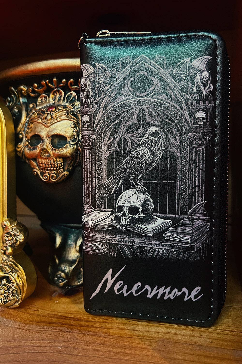 A black zip-around wallet with a detailed white gothic illustration of a raven, skull, books, and arches, with the word 'Nevermore' in white script at the bottom, displayed on a wooden surface next to a decorative skull object.