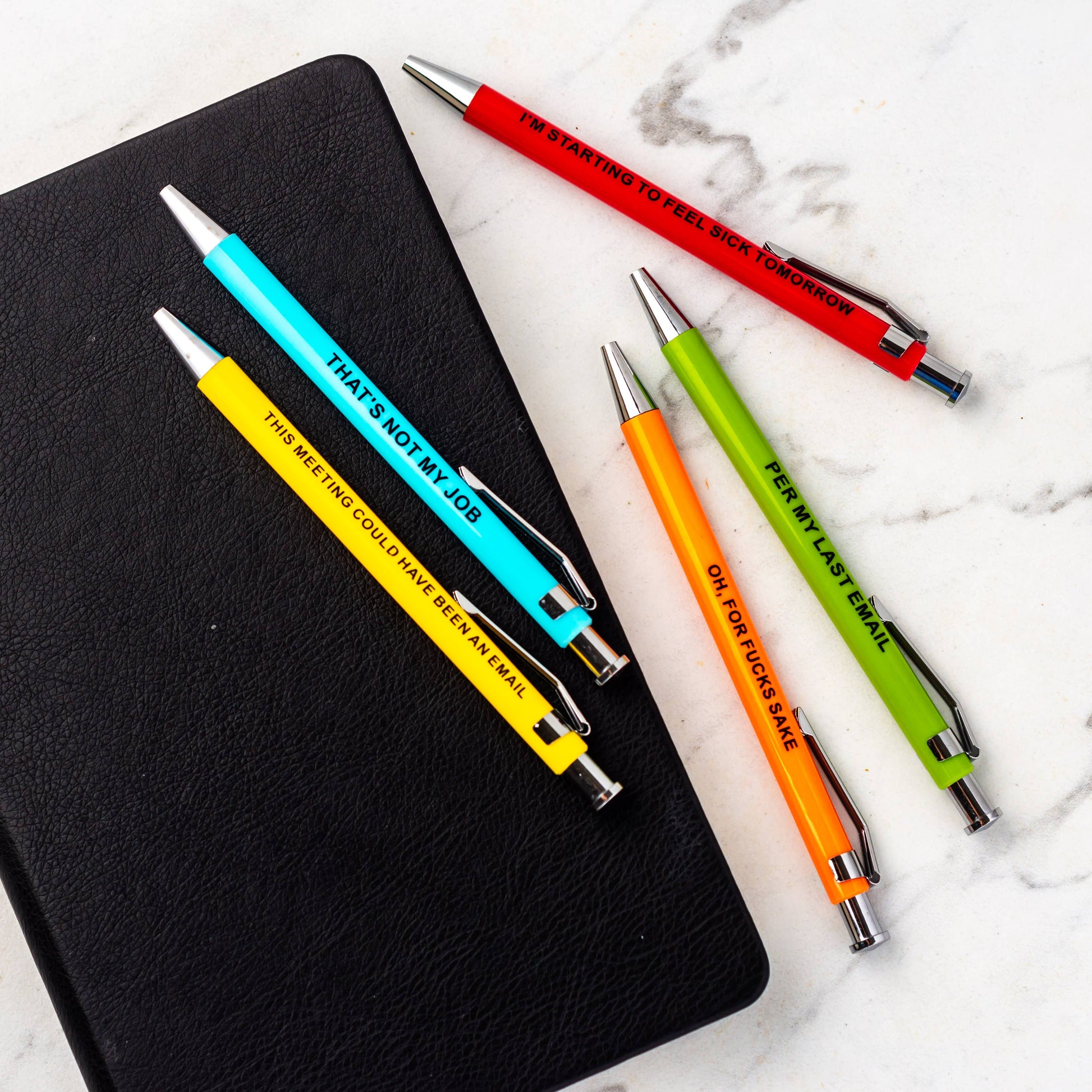 Colorful pens with humorous text on a black notebook and marble surface