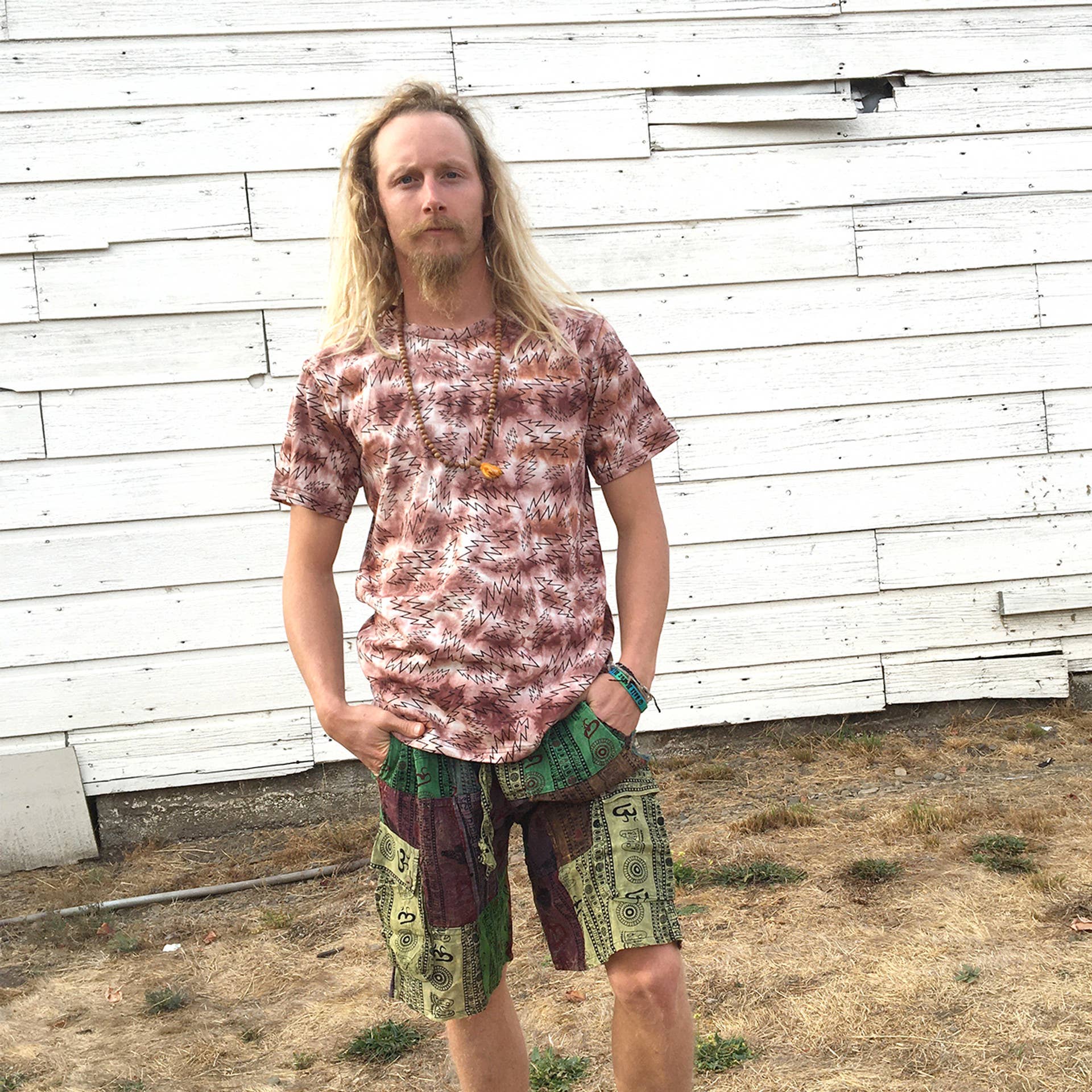 Man wearing a tie-dye shirt and colorful shorts standing in front of a white wooden wall.