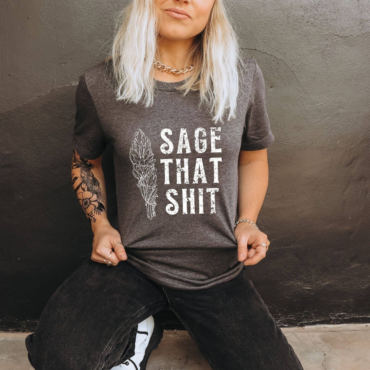 Person wearing a brown t-shirt with 'SAGE THAT SHIT' text and feather graphic against a dark background