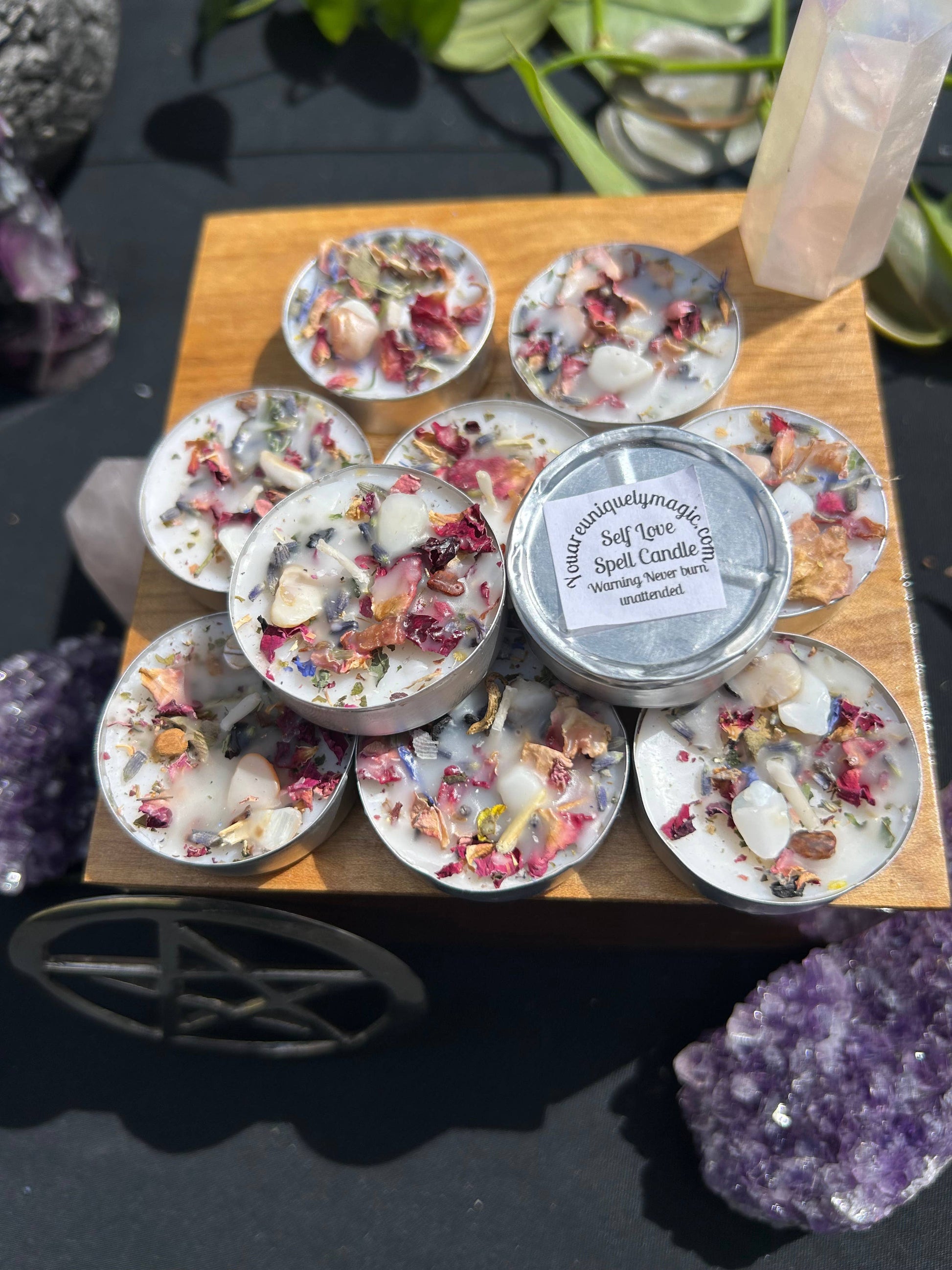 Small tins of decorative candles with floral elements on a wooden stand, surrounded by crystals and plants.