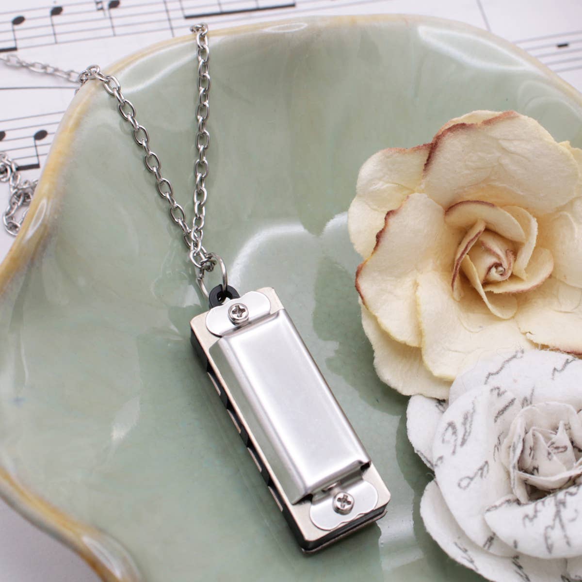 Metallic harmonica pendant on a chain with a white rose and decorative stone on a light green dish.