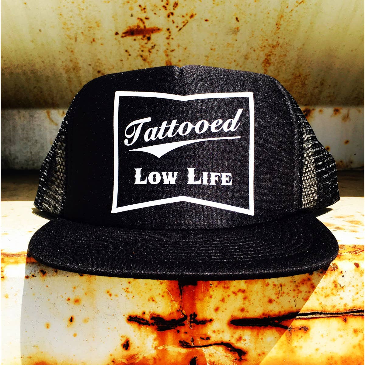 Black trucker hat with 'Tattooed Low Life' text on a rusty metal background