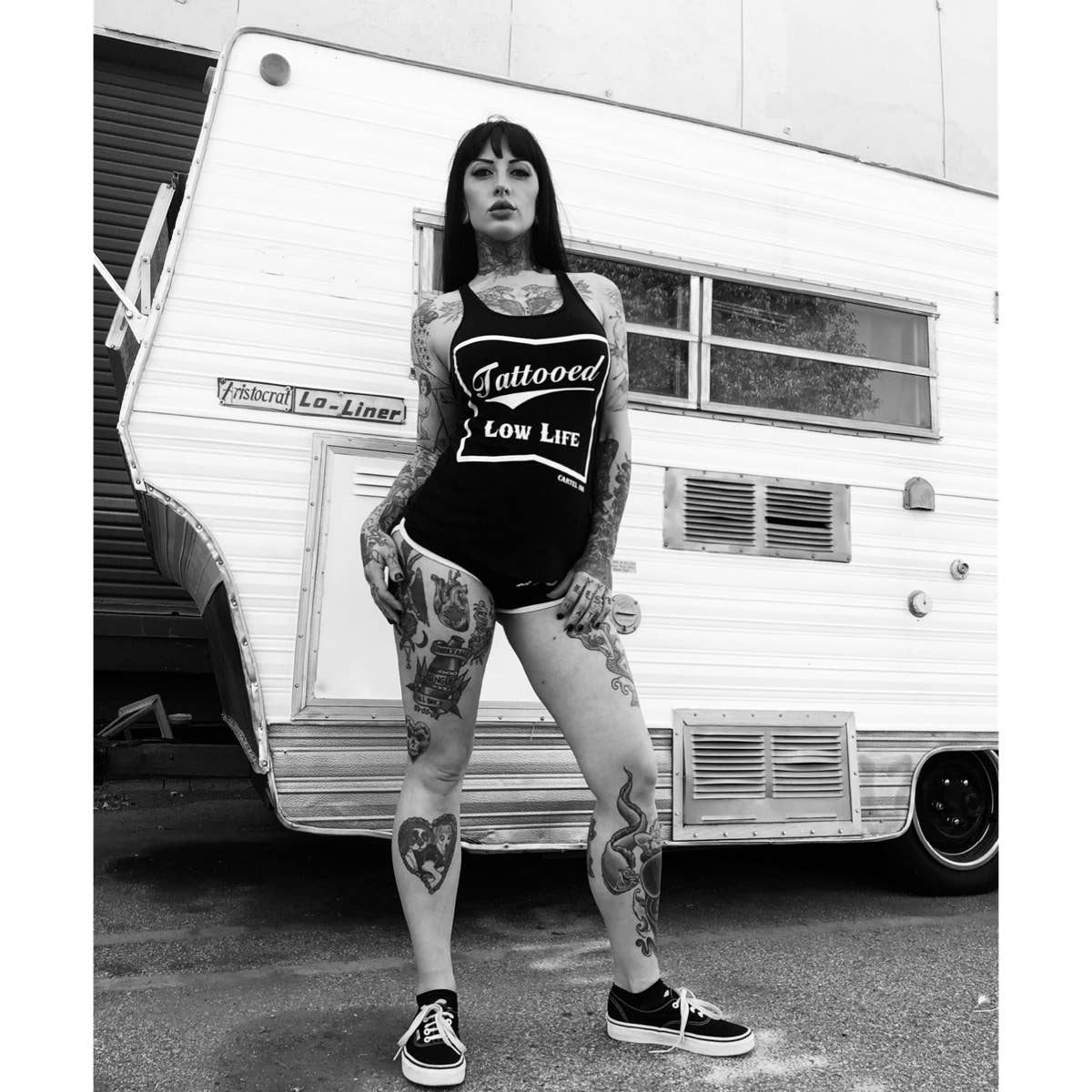 Person wearing a 'Tattooed Low Life' tank top standing in front of a vintage trailer.
