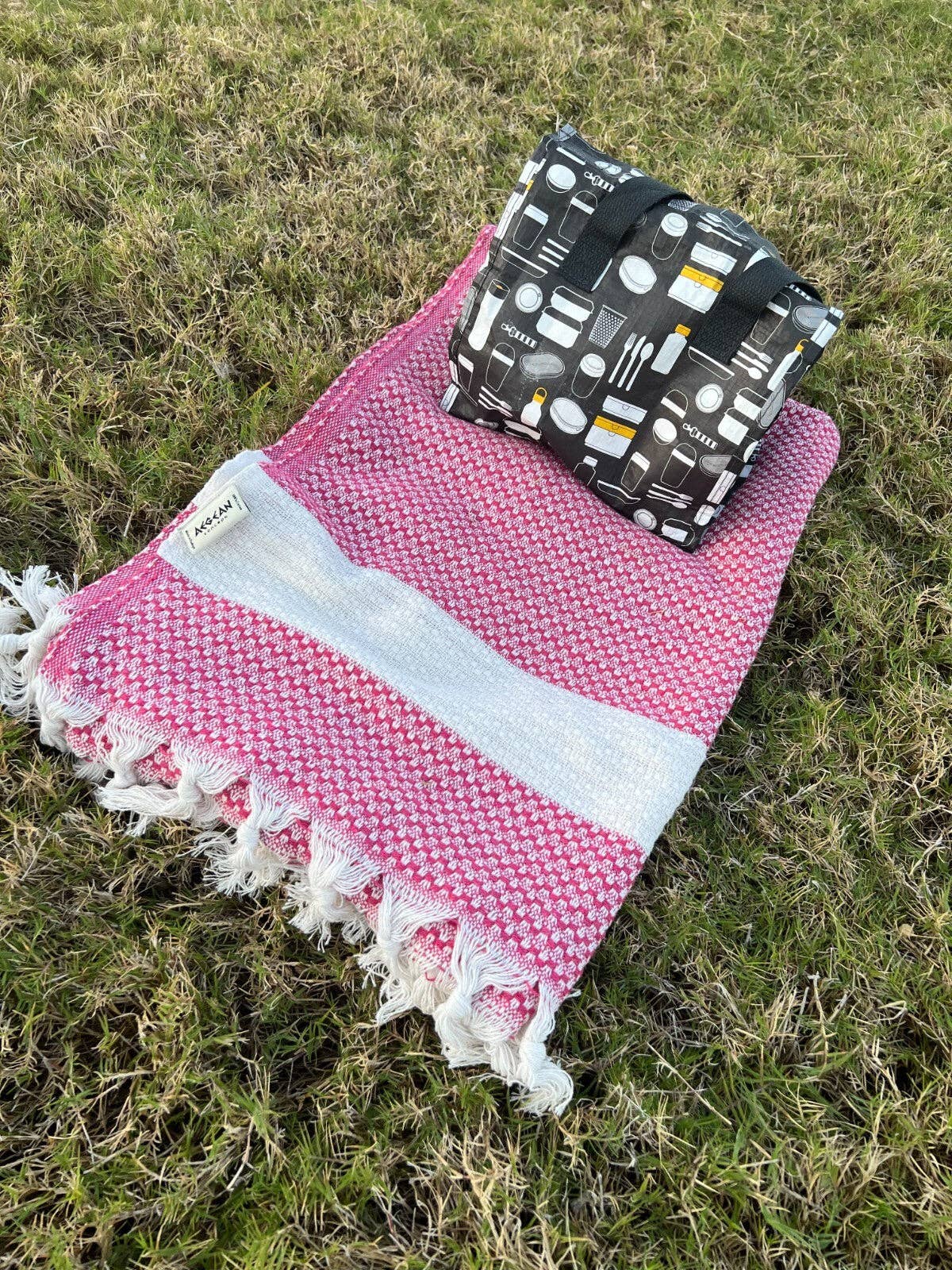 Pink towel with white fringe and black patterned bag on grass