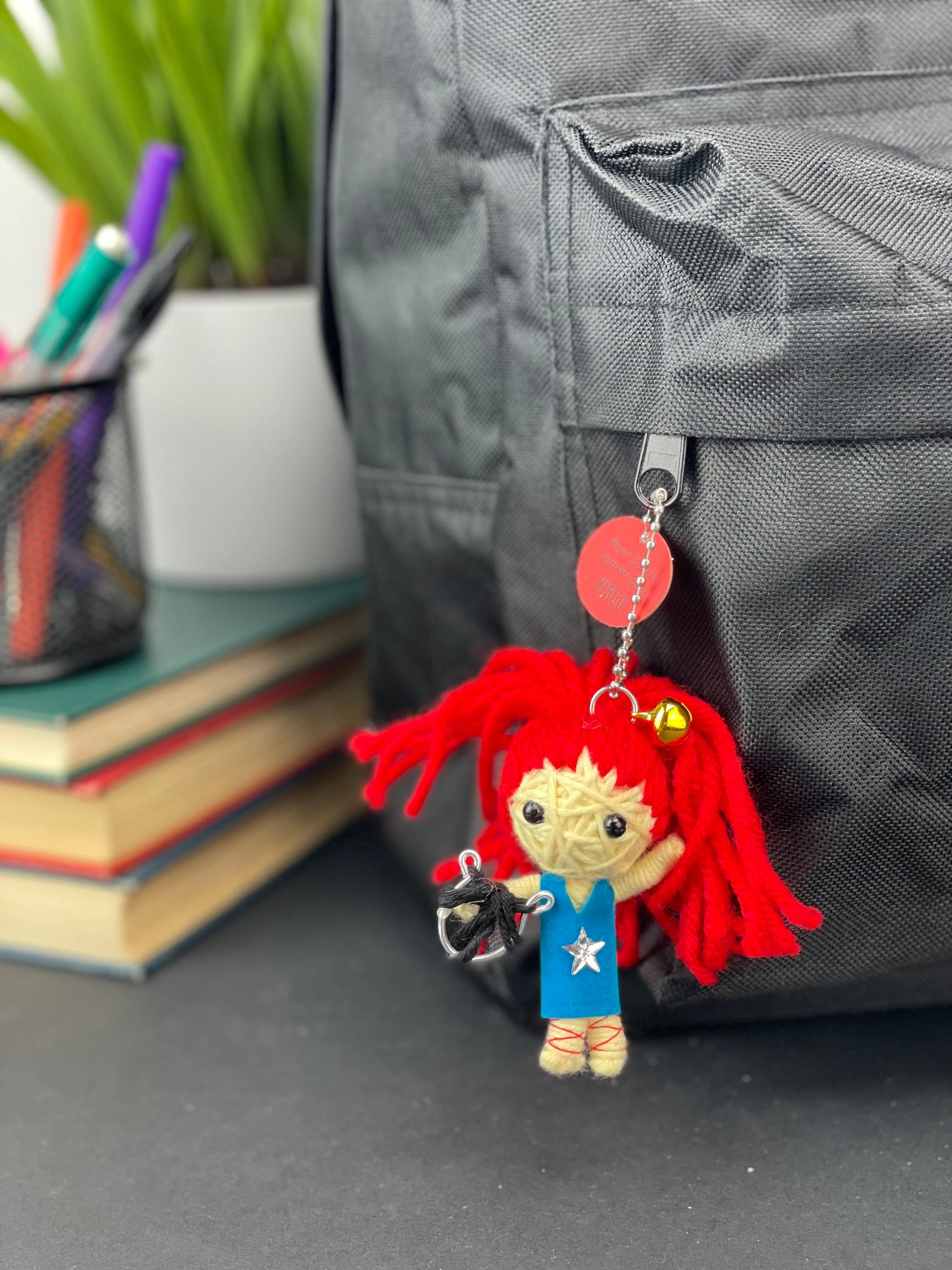 Keychain with red hair and blue outfit attached to a backpack, with books and stationery items in the background.