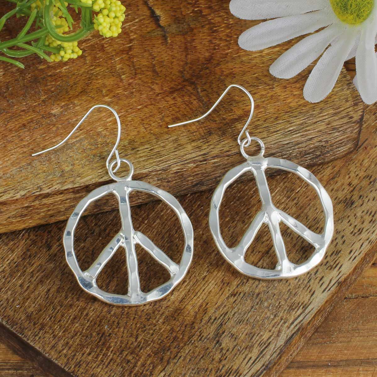 Silver peace sign earrings on a wooden surface with flowers.