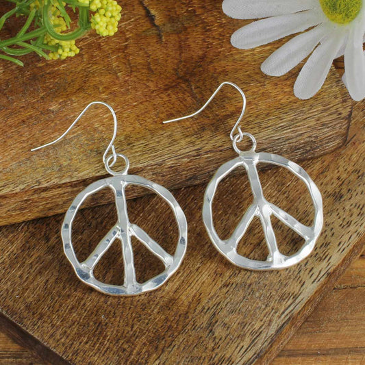 Silver peace sign earrings on a wooden surface with flowers.