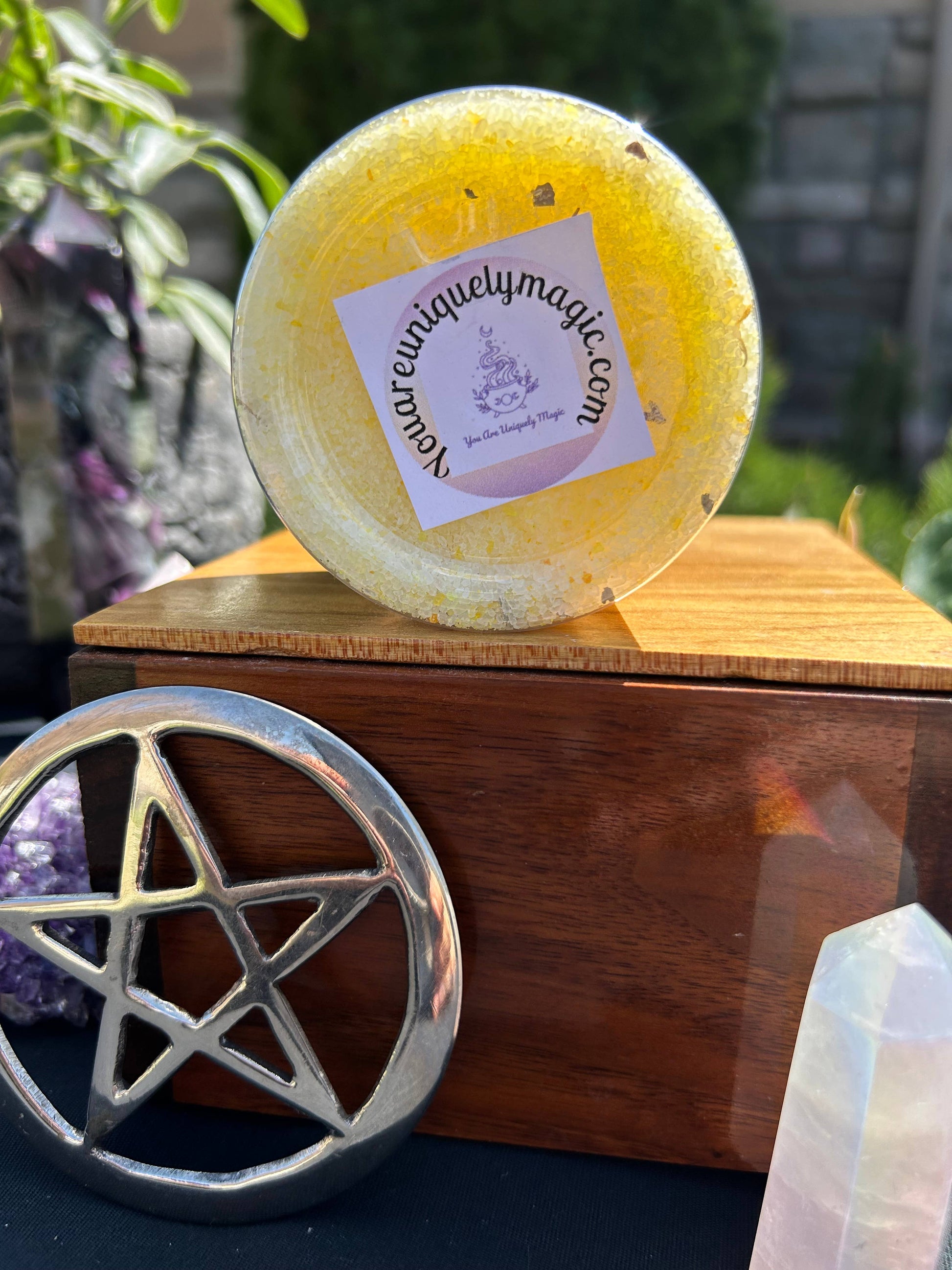Yellow bath bomb with a brand label on a wooden box, surrounded by crystals and a pentacle symbol.